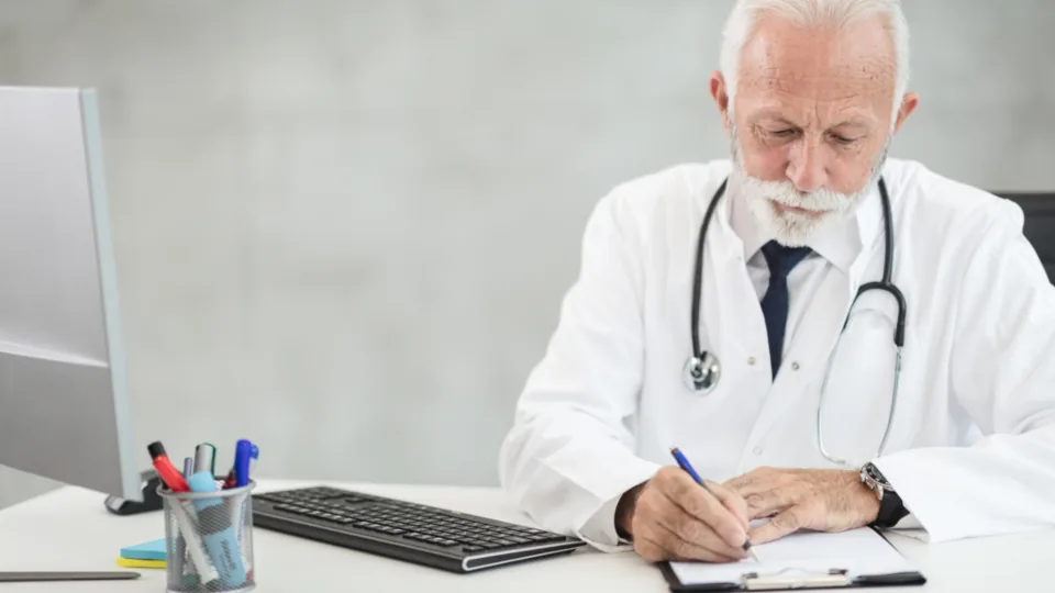 Médico de jaleco escrevendo à mesa do consultório, ao lado do computador