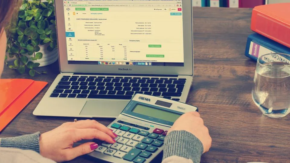 Mãos operando uma calculadora ao lado de um notebook sobre a mesa